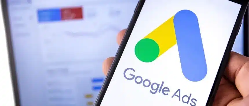 Móvil mostrando logo Google Ads con estadísticas detrás en portátil.
