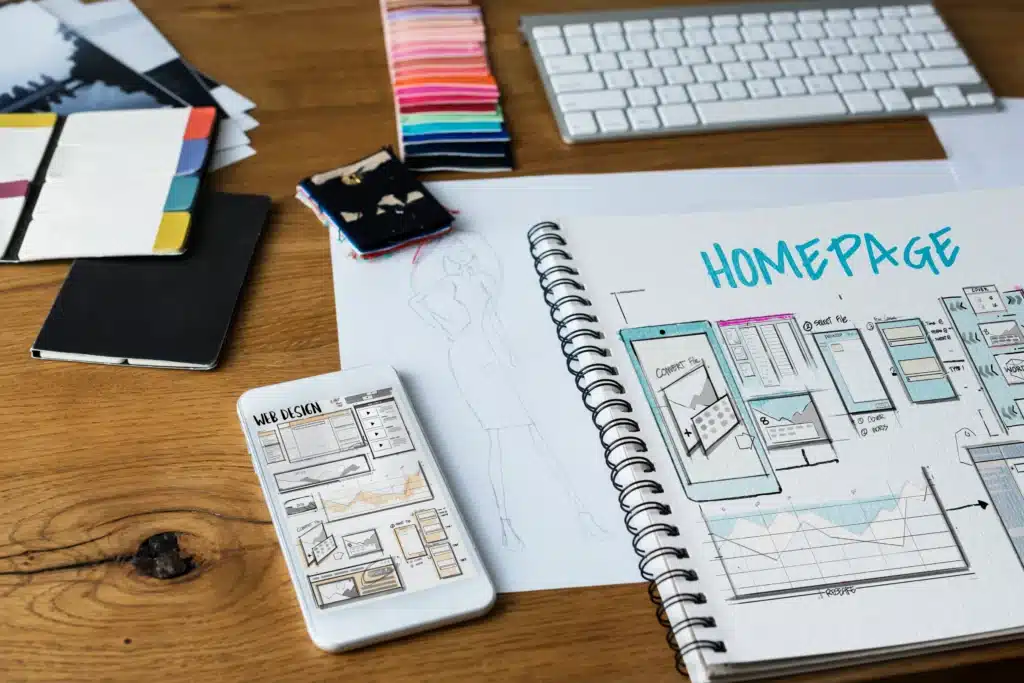Cuaderno abierto con bocetos de diseño web, esquemas de páginas y prototipos de una página de inicio, acompañado de muestras de colores, un teclado y un dispositivo móvil.