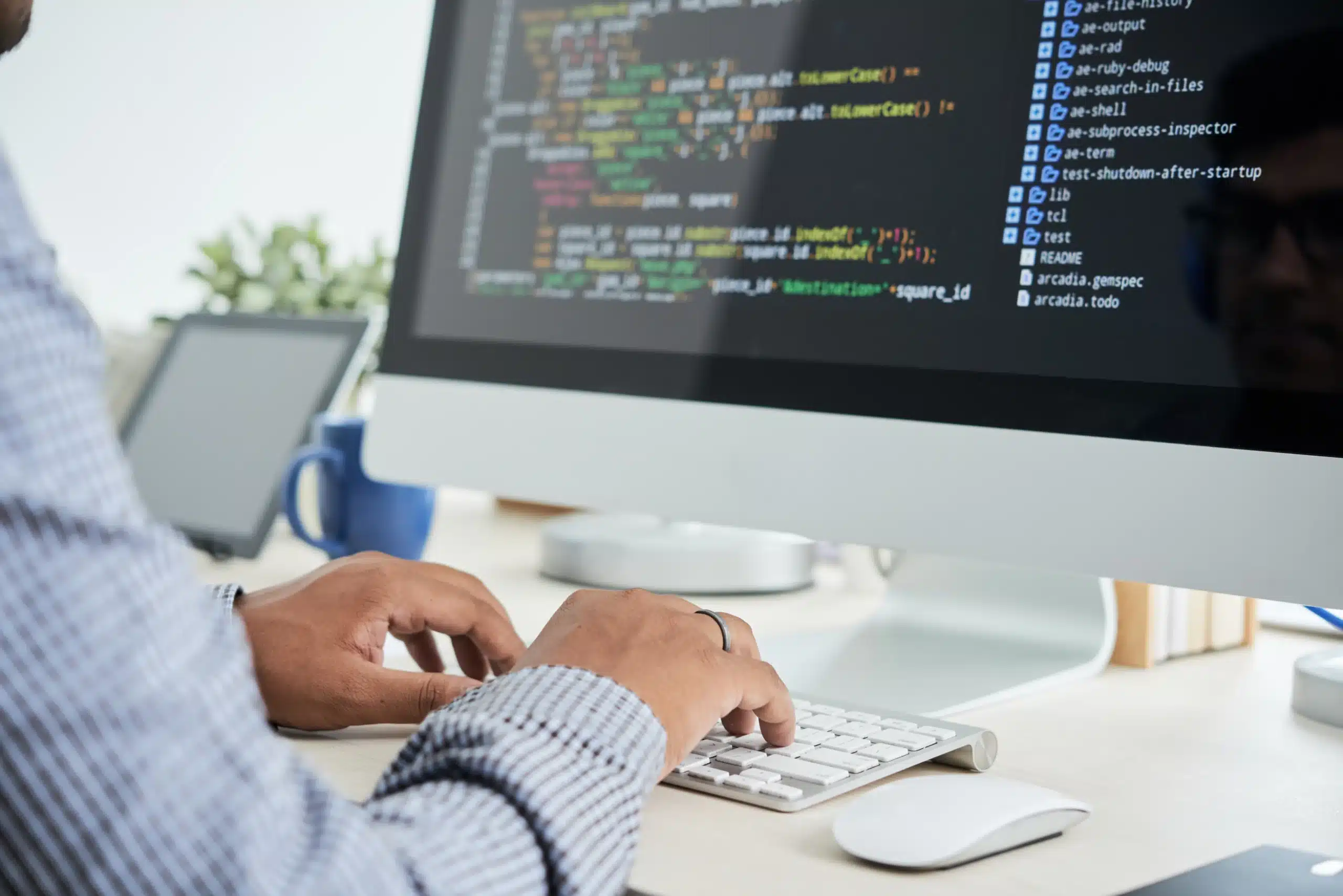 Persona trabajando con un teclado frente a un ordenador de sobremesa que muestra código de programación en la pantalla.