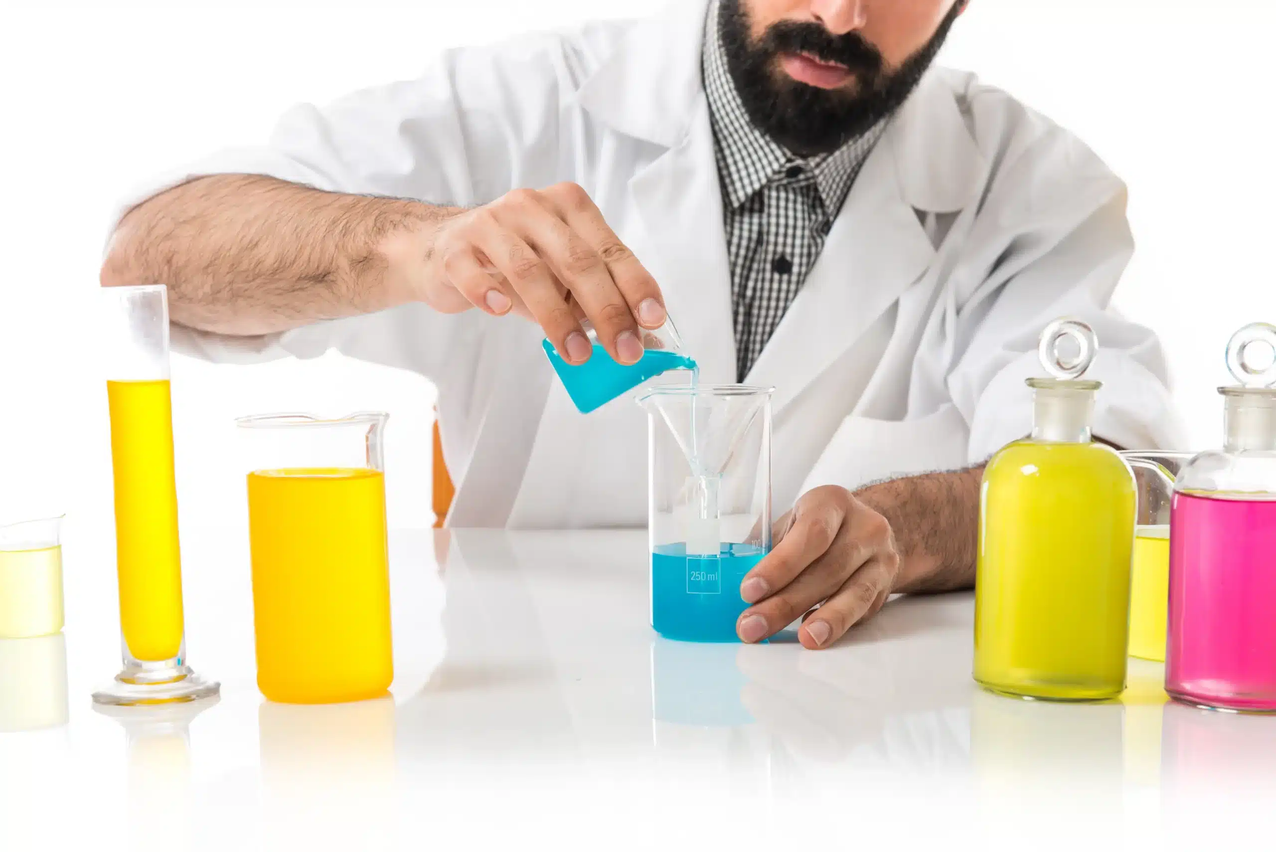 Productos químicos: 7 claves para almacenarlos y gestionarlos con seguridad 3 Profesional con bata blanca vierte un líquido azul en un recipiente de laboratorio junto a varios recipientes con líquidos de colores.